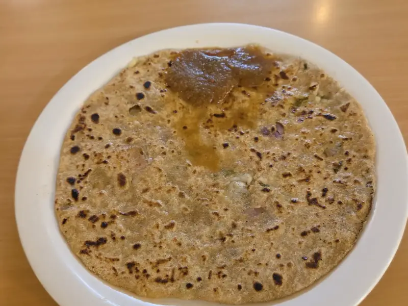 A cooked aloo paratha on a plate, browned in patches, with a bowl of curd beside it.