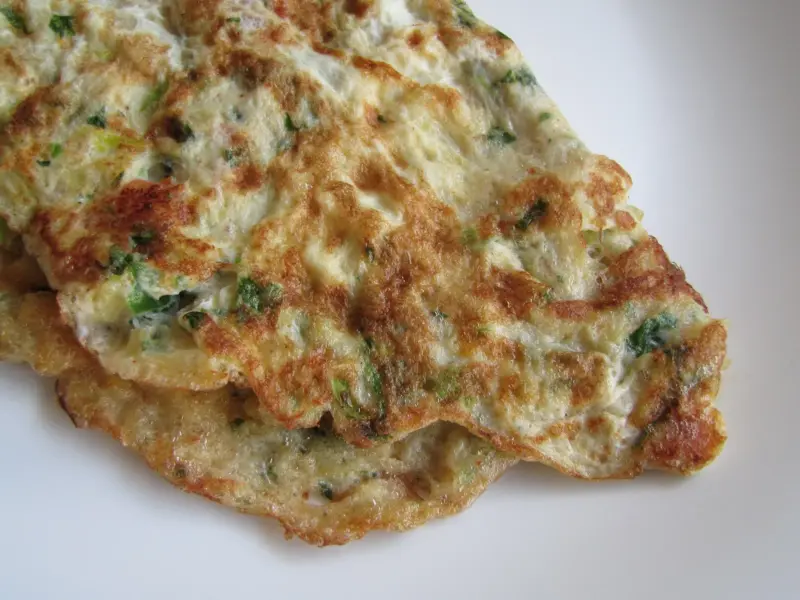 A folded masala omelette on a plate, flecked with onion, chilli and coriander.